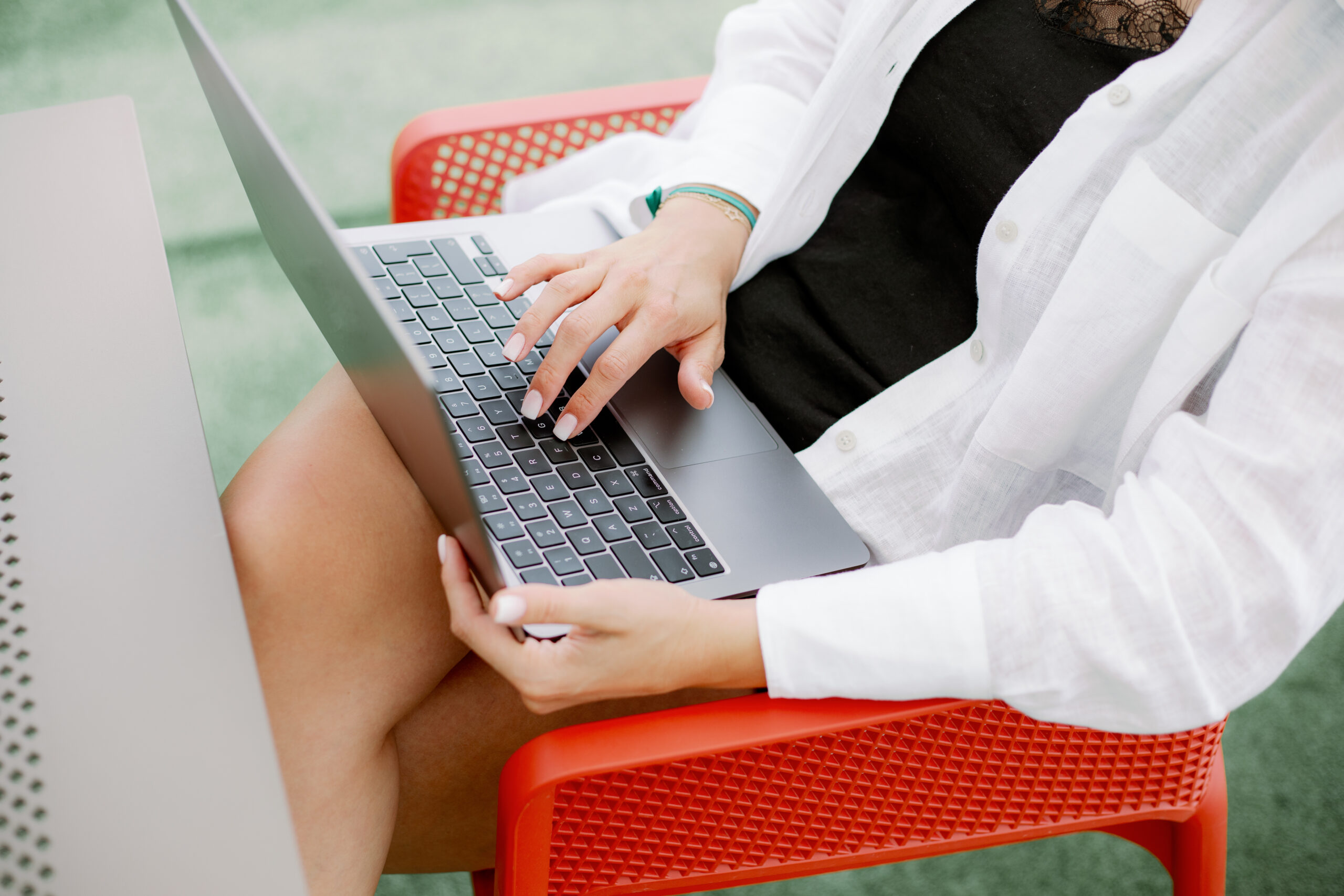 Close up of young woman in white shirt and black dress sitting on red chair with laptop on lap and typing using keyboard. Freelancer with delicate manicure and green bracelet busy with work.